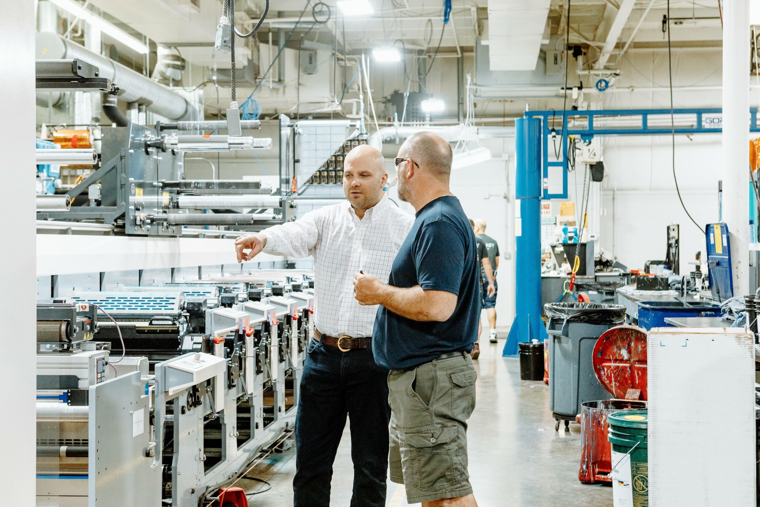 Two people stand in the Steinhauser pressroom in front a a flexographic printing press where custom labels for consumer packaged goods are printed.