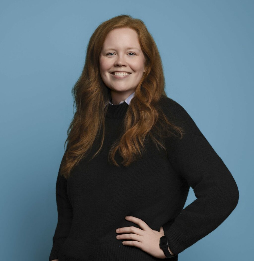 Woman stands posed with a hand on her hip smiling in front of a light blue background