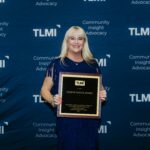 Tara Halpin, Steinhauser CEO, stands holding the 2025 Eugene Singer Award with a TLMI step and repeat behind her.